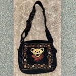 Black messenger bag with an embroidered golden bear face and decorative floral border design.