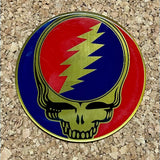 A circular, enamel-coated metal badge featuring the iconic grateful dead skull design with a red and blue background and gold lightning bolt.