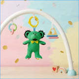 A green plush bear baby toy with black hair and a yellow bow tie hangs from a white baby gym arch. The arch has other hanging toys, including a pastel ring and a triangle rattle.