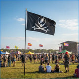 Festival scene with a large flag featuring a skull and crossbones design, people milling about, and a stage in the background.