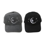 Two baseball caps, one in dark gray and the other black, both featuring a white embroidered design of a crescent moon with a face and small stars.